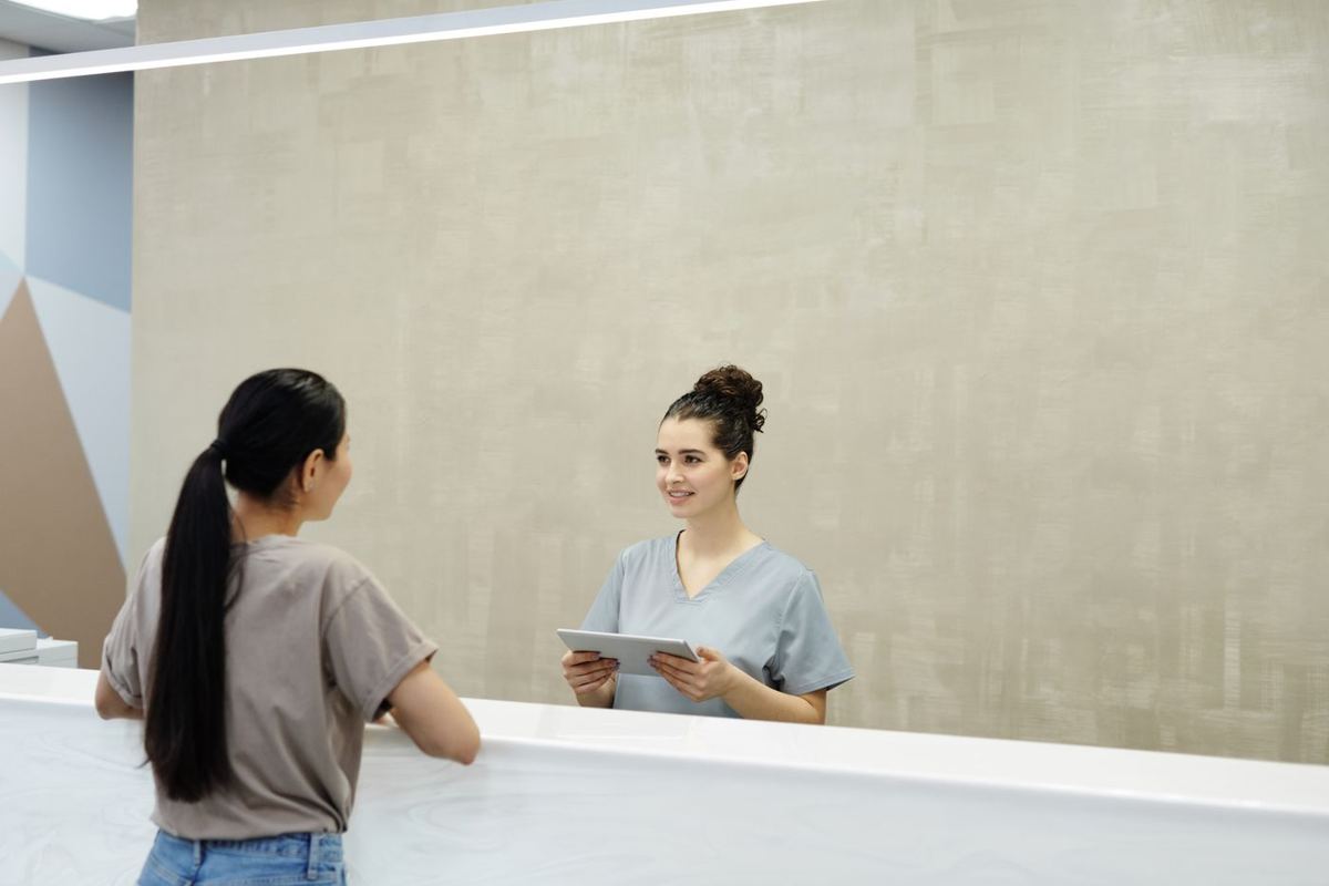A smiling clinic receptionist with a tablet welcomes a patient at the front desk